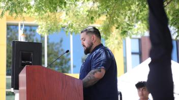 A bearded man in a navy shirt with a visible tattoo addresses the audience from the podium during the UC Davis Memorial Day Ceremony 2025