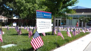 A field of small American flags and a sign honoring 136 fallen Aggies is displayed on a grassy lawn outside a campus building during the UC Davis Memorial Day Ceremony 2025