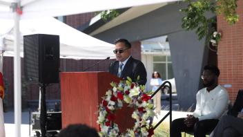 A man in a suit and sunglasses speaks at a podium beside a floral wreath, with seated guests and onlookers behind him, during the UC Davis Memorial Day Ceremony 2025