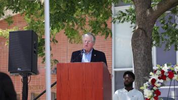 A man speaks at a podium beneath a large tree while seated guests, including a floral wreath, listen during the UC Davis Memorial Day Ceremony 2025