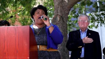 A woman sings passionately into a microphone at a podium while a man stands beside her with his hand over his heart during the UC Davis Memorial Day Ceremony 2025