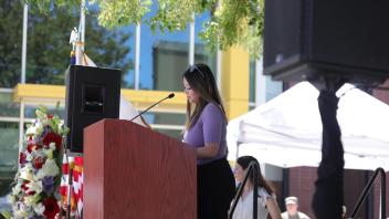 A woman stands at a podium reading into a microphone, with a floral wreath and American flags nearby, during the UC Davis Memorial Day Ceremony 