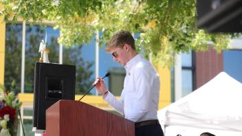 A young man in sunglasses reads from a paper at the podium under tree shade