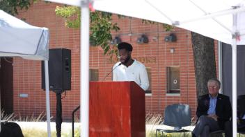 A young man stands at a podium reading to the audience while seated speakers listen nearby, beneath a white canopy