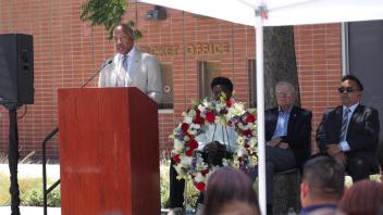 Chancellor Gary S. May addresses the audience from a podium as panelists and attendees, seated under a canopy, observe a large ceremonial wreath