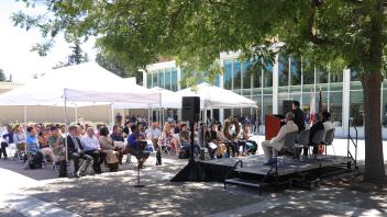 Dozens of attendees sit under white canopies listening to speakers on stage during the UC Davis Memorial Day Ceremony 2025, held outside a modern campus building surrounded by trees.jpg