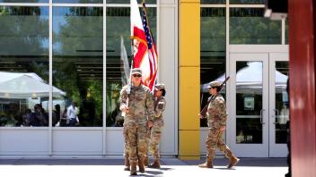 Four uniformed service members march in formation carrying the American and California flags, with reflections of the audience visible in the building’s glass behind them