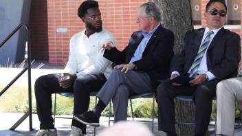 Three men sit on a stage under a tree at the UC Davis Memorial Day Ceremony 2025, with two engaged in conversation while the third looks ahead, holding a program
