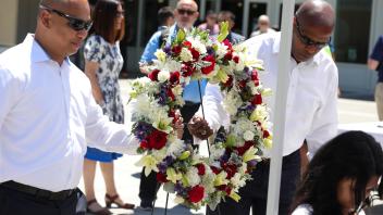 Two men in white dress shirts carefully carry a large floral wreath adorned with red, white, and purple flowers during the UC Davis Memorial Day Ceremony 2025, as people gather in the background