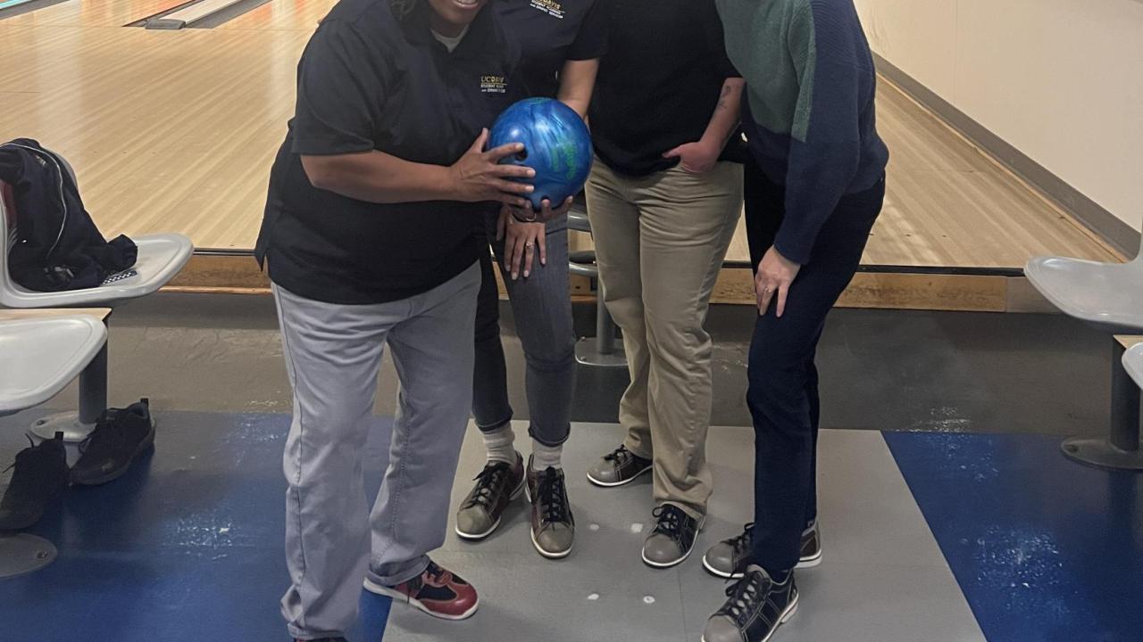 Four staff members smile and pose in front of the Games Area bowling lanes. One staff members holds up a blue bowling ball.