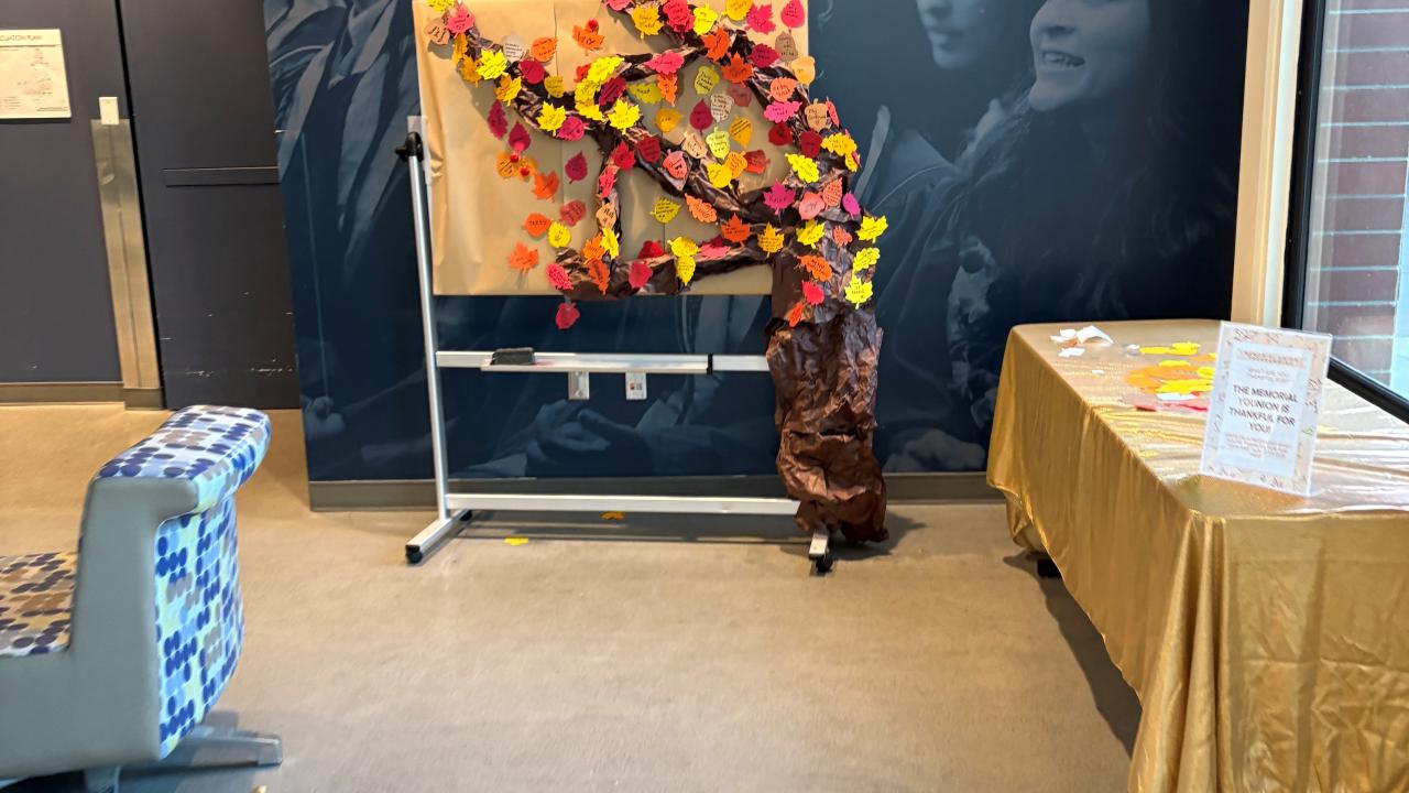 A display features a large paper tree attached to a whiteboard, with brown paper forming the trunk and colorful paper leaves in red, orange, yellow, and pink covering the branches. Each leaf appears to have handwritten messages. To the right, there’s a table covered in a gold tablecloth with more supplies for writing notes. The setup is in a bright indoor space.