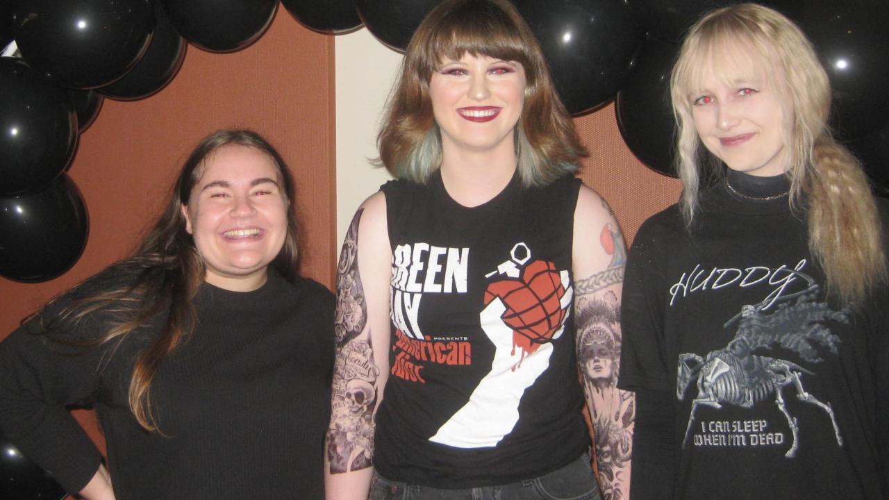 Three students dressed in Emo style attire in front of a balloon arch, posing for a picture.