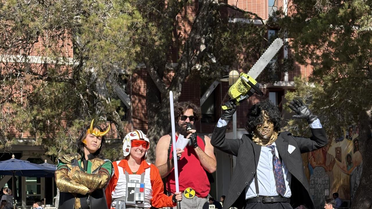 Four people in costumes stand on an outdoor stage decorated with inflatable pumpkins. From left to right: the first person is dressed as Loki with a green and gold outfit and horned helmet; the second is wearing an orange Rebel pilot uniform from Star Wars; the third is in a red tank top, gray pants, sunglasses, and holding a toy weapon resembling a bat with spikes; the fourth is dressed as Leatherface in a suit, tie, mask, and holding a chainsaw overhead. Trees, tables, and a brick building are in the back