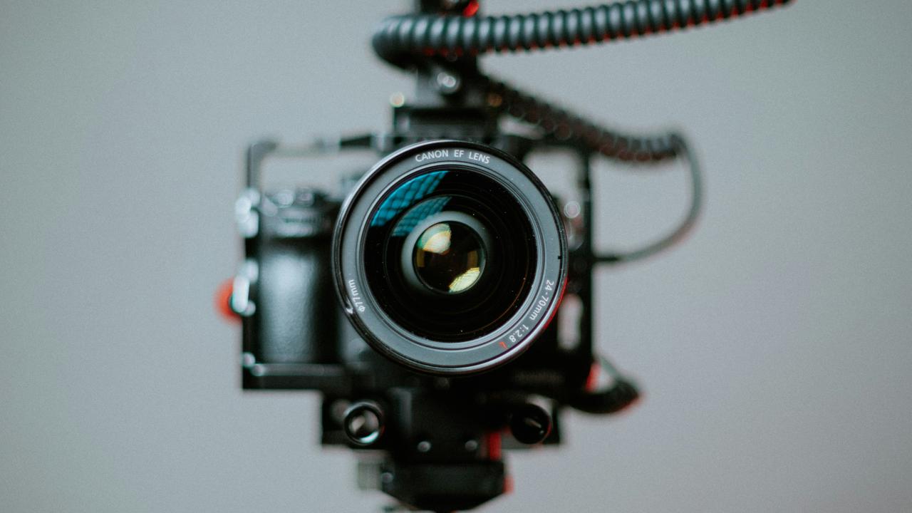 Close-up of a professional Canon EF camera lens mounted on a rig with coiled cables, focused directly at the viewer against a plain gray background.