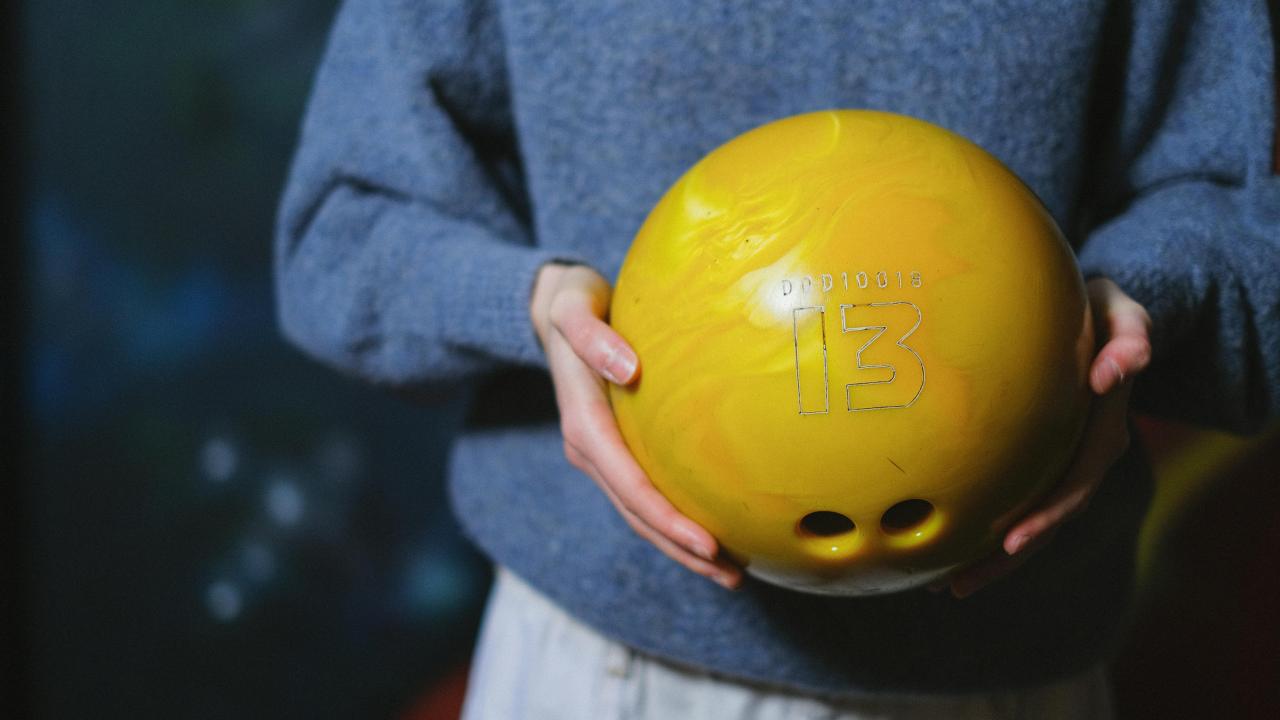Person in a dusty blue sweater and light great pants is holding a yellow bowling ball with the number 13 etched in a white online showing on the ball. The photo cuts off below their waist, so only one silver button on the pants are showing. 
