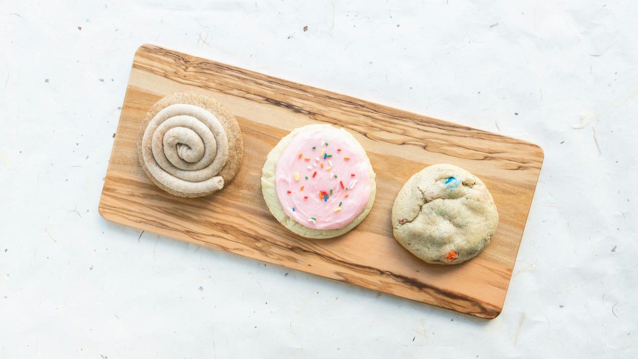 A wooden serving board holds three cookies in a row. From left to right: a cinnamon roll-style cookie with a spiral of frosting, a sugar cookie topped with pink frosting and rainbow sprinkles, and a thick cookie with colorful candy pieces baked inside.