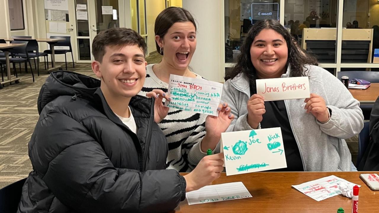 Three students hold up their team name at Trivia Night