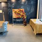 A display features a large paper tree attached to a whiteboard, with brown paper forming the trunk and colorful paper leaves in red, orange, yellow, and pink covering the branches. Each leaf appears to have handwritten messages. To the right, there’s a table covered in a gold tablecloth with more supplies for writing notes. The setup is in a bright indoor space.