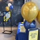 A back-to-school photo station set up in a campus building, decorated with gold, blue, and white balloons. A table with a blue tablecloth holds props labeled with different school years. Signs encourage students to take their "first day of school" photo, with one sign reading “Take Your First Day of School Photo!” and another advertising “It’s my First Year!” In the background, a wall mural shows graduates in caps and gowns.