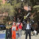 Four people in costumes stand on an outdoor stage decorated with inflatable pumpkins. From left to right: the first person is dressed as Loki with a green and gold outfit and horned helmet; the second is wearing an orange Rebel pilot uniform from Star Wars; the third is in a red tank top, gray pants, sunglasses, and holding a toy weapon resembling a bat with spikes; the fourth is dressed as Leatherface in a suit, tie, mask, and holding a chainsaw overhead. Trees, tables, and a brick building are in the back
