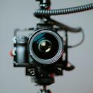 Close-up of a professional Canon EF camera lens mounted on a rig with coiled cables, focused directly at the viewer against a plain gray background.