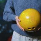 Person in a dusty blue sweater and light great pants is holding a yellow bowling ball with the number 13 etched in a white online showing on the ball. The photo cuts off below their waist, so only one silver button on the pants are showing. 