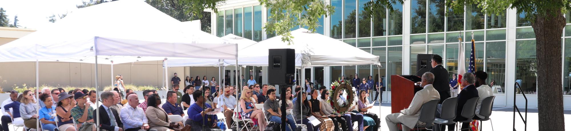 Dozens of attendees sit under white canopies listening to speakers on stage during the UC Davis Memorial Day Ceremony 2025, held outside a modern campus building surrounded by trees.jpg